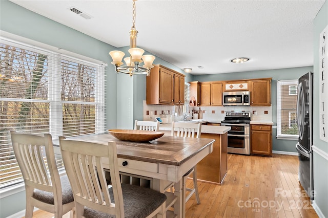 kitchen with a chandelier, appliances with stainless steel finishes, pendant lighting, light hardwood / wood-style floors, and decorative backsplash