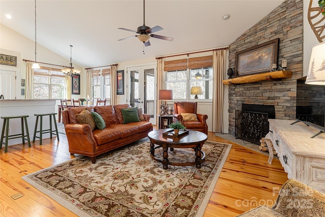 living room featuring a stone fireplace, ceiling fan with notable chandelier, high vaulted ceiling, light wood-type flooring, and french doors