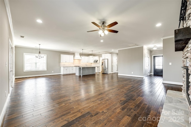 unfurnished living room with a stone fireplace, ceiling fan with notable chandelier, visible vents, ornamental molding, and dark wood-style floors