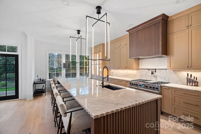 kitchen with sink, tasteful backsplash, a center island with sink, decorative light fixtures, and wall chimney exhaust hood