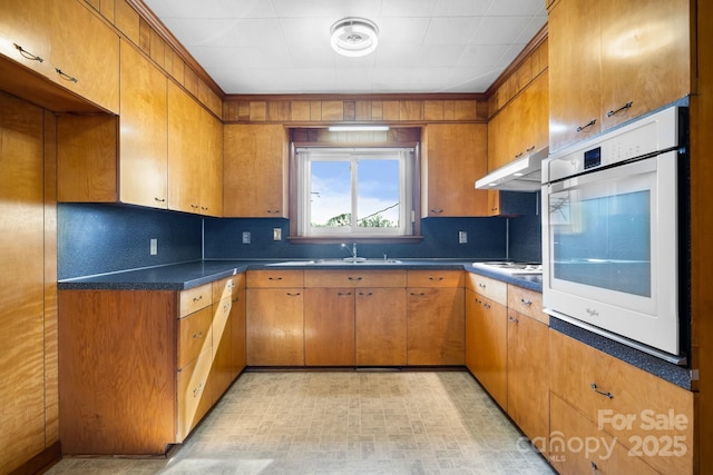 kitchen with brown cabinetry, dark countertops, white appliances, and under cabinet range hood