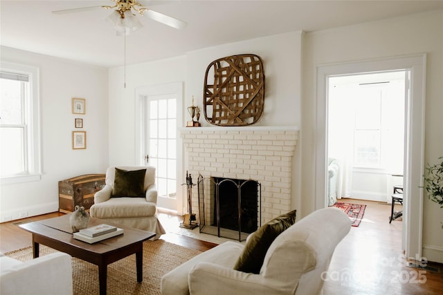 living room with a brick fireplace, ceiling fan, baseboards, and wood finished floors