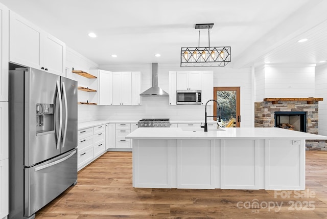 kitchen with wall chimney range hood, hanging light fixtures, stainless steel appliances, and an island with sink