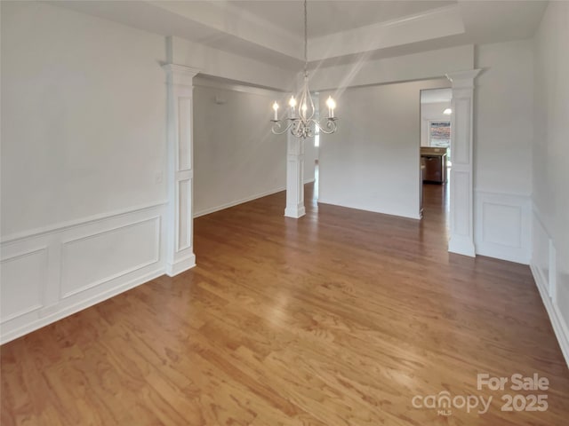 unfurnished dining area with an inviting chandelier, a tray ceiling, wood-type flooring, and ornate columns