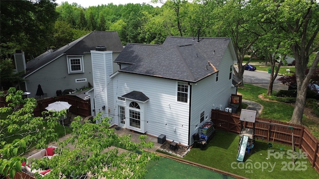 back of house with a wooded view, a chimney, a lawn, a shingled roof, and fence