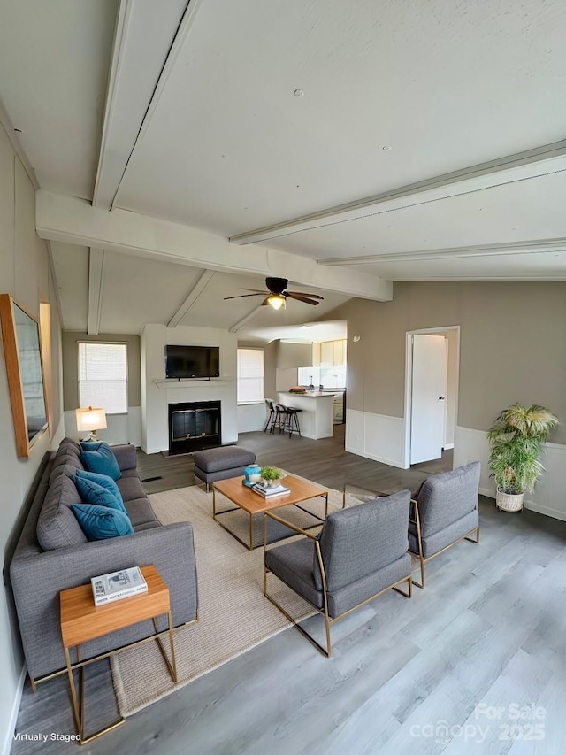 living area featuring a wainscoted wall, lofted ceiling with beams, a glass covered fireplace, ceiling fan, and wood finished floors