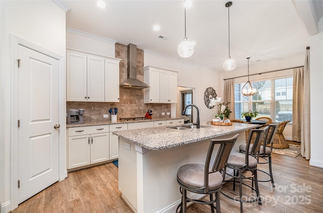 kitchen featuring an island with sink, sink, white cabinets, hanging light fixtures, and wall chimney range hood