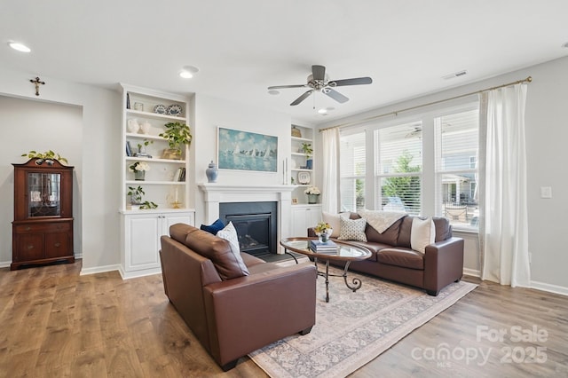 living area with wood finished floors, a glass covered fireplace, ceiling fan, baseboards, and visible vents