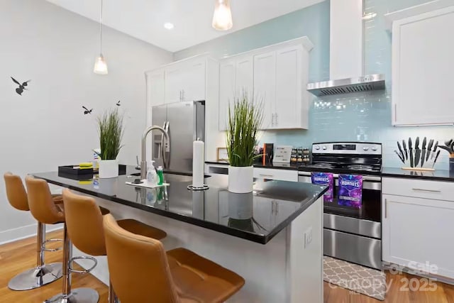 kitchen with a breakfast bar, wall chimney range hood, an island with sink, stainless steel appliances, and white cabinets