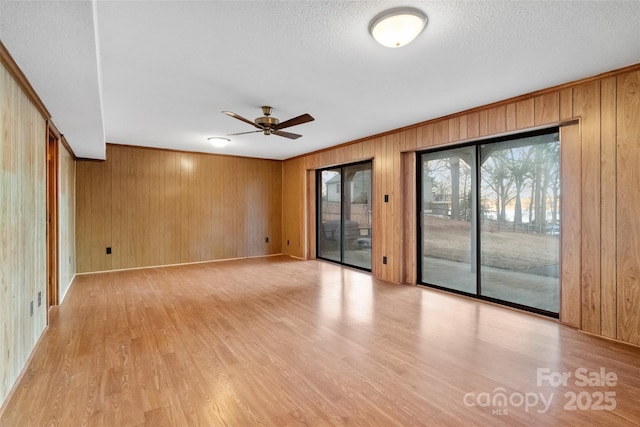 empty room with a textured ceiling, light wood-type flooring, ornamental molding, wooden walls, and ceiling fan