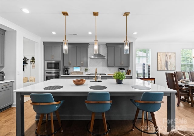 kitchen with a center island with sink, appliances with stainless steel finishes, gray cabinetry, and wall chimney range hood