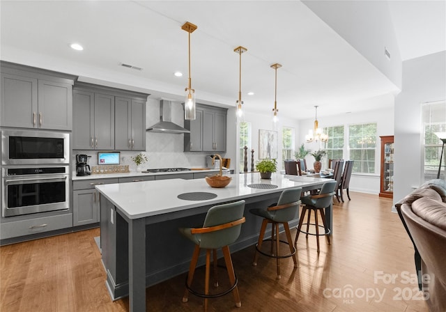 kitchen with a kitchen island with sink, gray cabinets, wall chimney exhaust hood, and appliances with stainless steel finishes