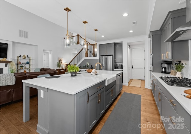 kitchen featuring gray cabinetry, hanging light fixtures, a kitchen island with sink, stainless steel appliances, and wall chimney exhaust hood