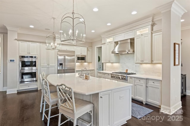 kitchen featuring wall chimney exhaust hood, white cabinetry, built in appliances, hanging light fixtures, and an island with sink