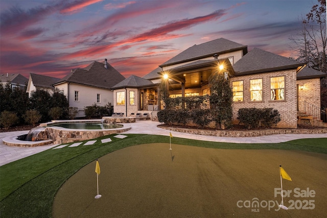 back of house at dusk featuring brick siding, concrete driveway, a patio area, an in ground hot tub, and an outdoor pool