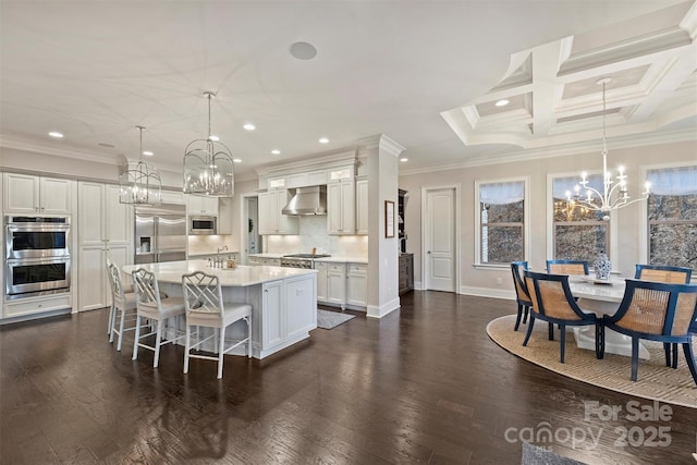 kitchen with built in appliances, a notable chandelier, dark wood-style flooring, light countertops, and wall chimney range hood