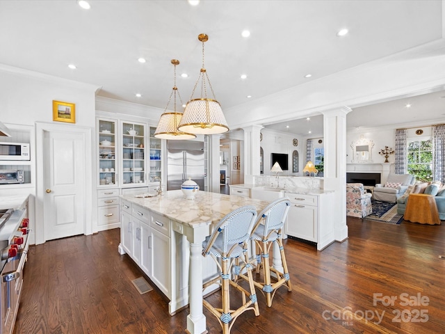 kitchen featuring high quality appliances, open floor plan, decorative columns, a large island with sink, and glass insert cabinets
