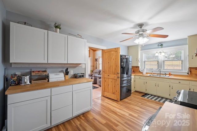 kitchen with sink, butcher block counters, white cabinets, black refrigerator with ice dispenser, and light wood-type flooring