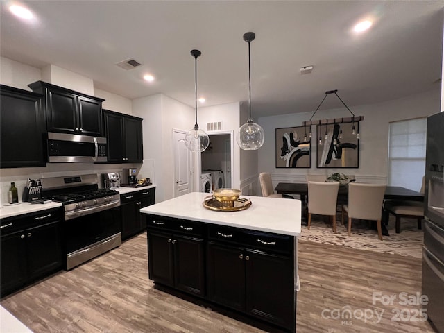 kitchen featuring pendant lighting, washer and clothes dryer, stainless steel appliances, light hardwood / wood-style floors, and an island with sink