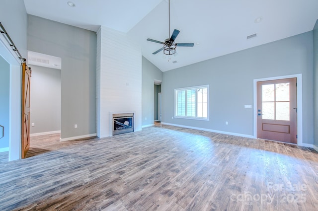 unfurnished living room featuring high vaulted ceiling, a large fireplace, hardwood / wood-style flooring, ceiling fan, and a barn door