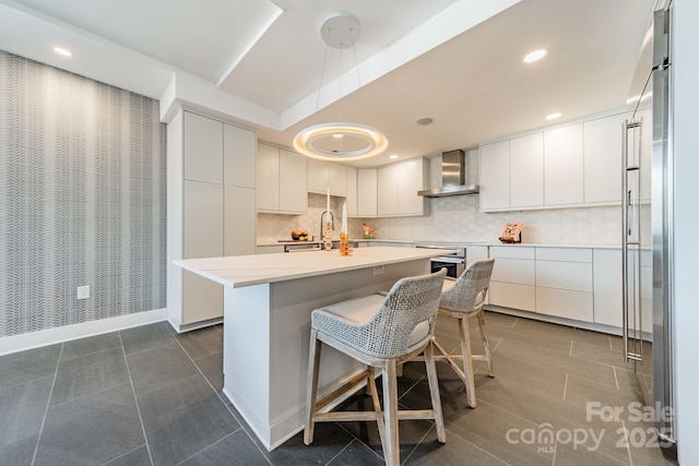 kitchen featuring backsplash, white cabinetry, pendant lighting, wall chimney range hood, and an island with sink