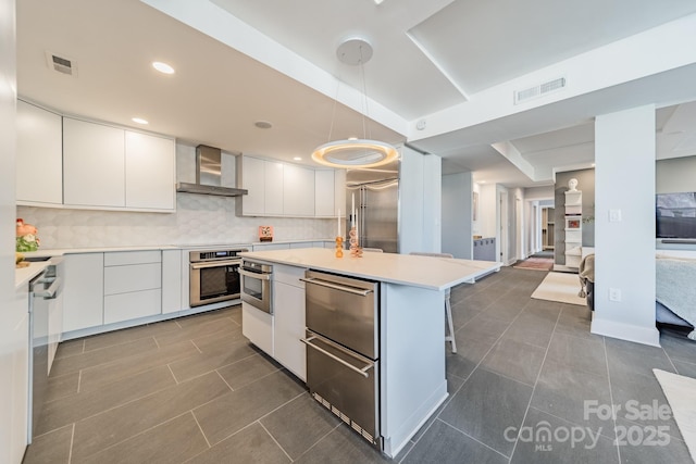 kitchen with appliances with stainless steel finishes, wall chimney exhaust hood, pendant lighting, a center island, and white cabinetry