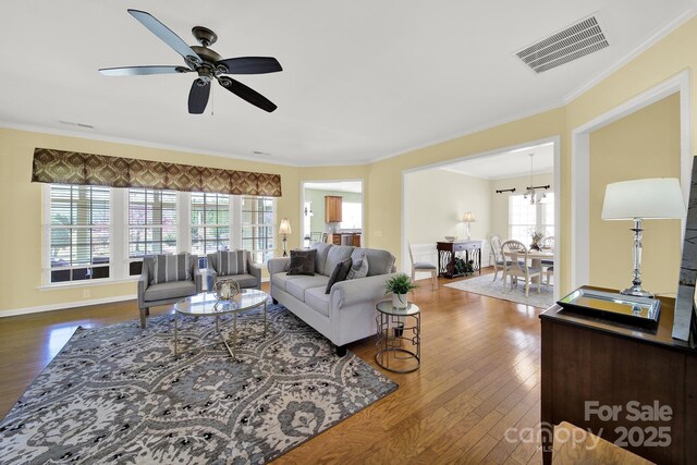 living area with crown molding, ceiling fan with notable chandelier, baseboards, dark wood finished floors, and visible vents
