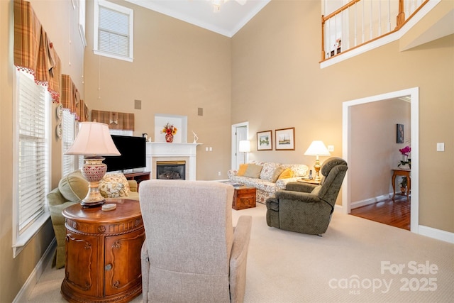 living area featuring baseboards, a glass covered fireplace, ornamental molding, a high ceiling, and carpet floors