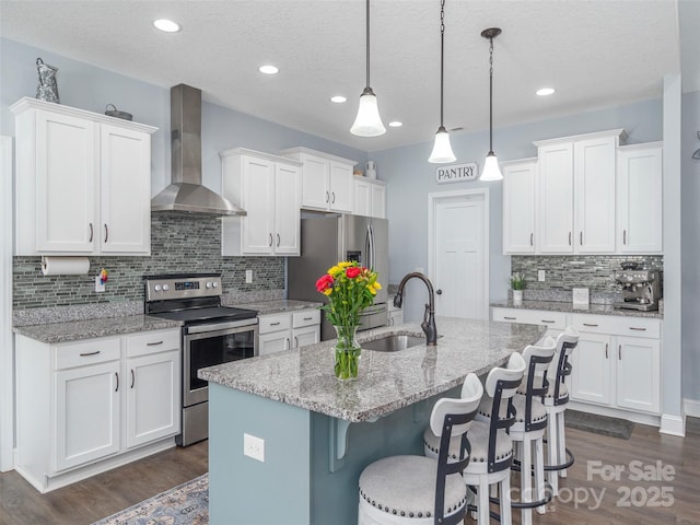 kitchen featuring wall chimney exhaust hood, sink, hanging light fixtures, appliances with stainless steel finishes, and white cabinets