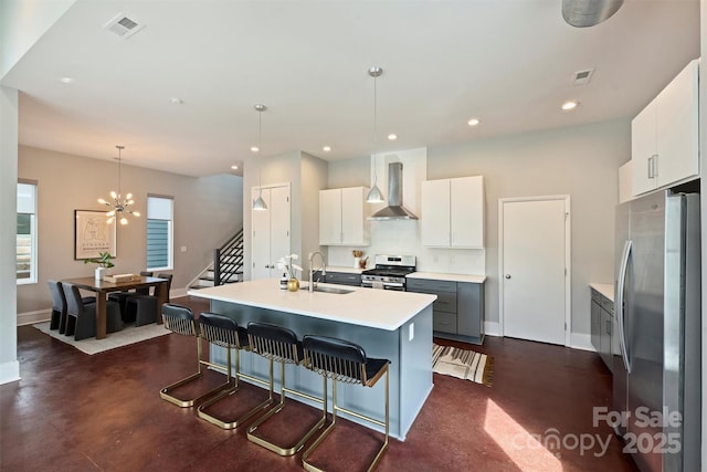 kitchen with wall chimney range hood, appliances with stainless steel finishes, a kitchen island with sink, hanging light fixtures, and white cabinets