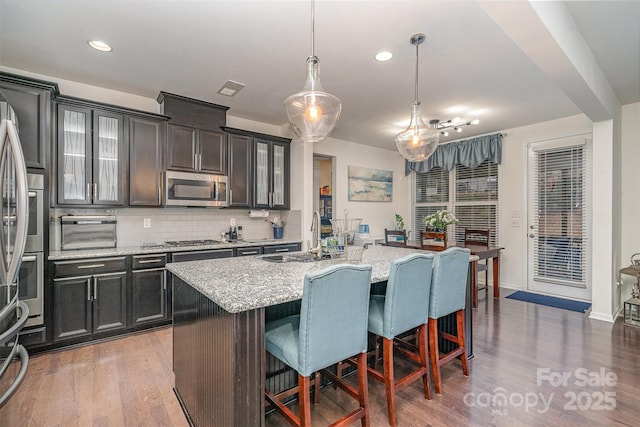 kitchen with sink, hanging light fixtures, backsplash, stainless steel appliances, and an island with sink