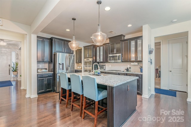 kitchen with stainless steel appliances, a kitchen breakfast bar, light stone countertops, an island with sink, and decorative light fixtures