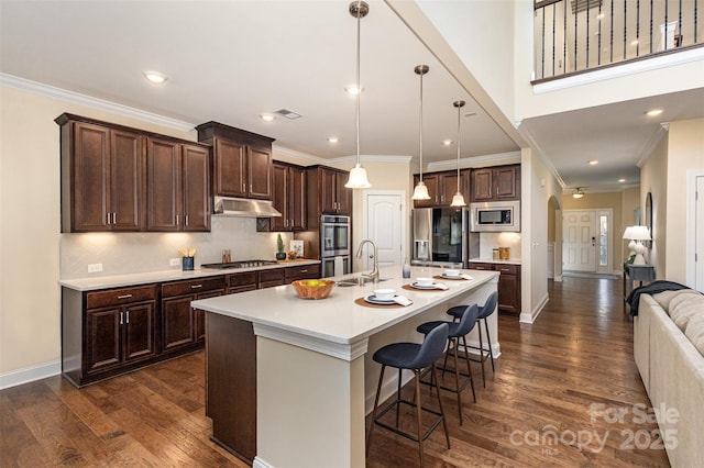 kitchen featuring arched walkways, dark brown cabinetry, under cabinet range hood, stainless steel appliances, and open floor plan