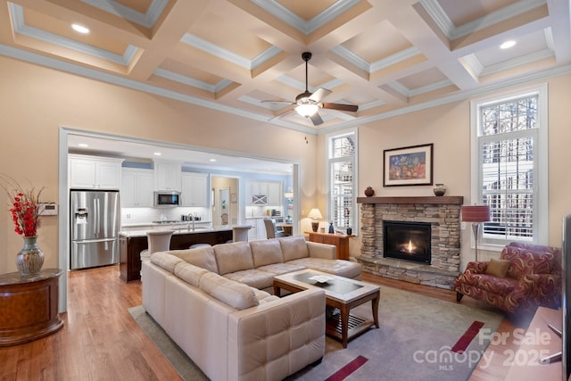 living room with ceiling fan, light hardwood / wood-style flooring, beamed ceiling, and a stone fireplace