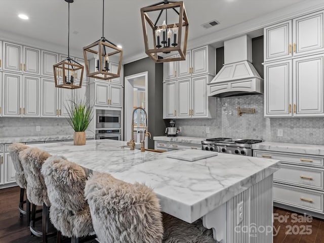 kitchen with visible vents, a kitchen island with sink, custom range hood, appliances with stainless steel finishes, and a notable chandelier