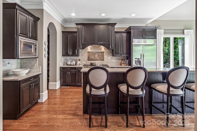 kitchen with dark hardwood / wood-style floors, built in appliances, dark brown cabinetry, light stone countertops, and an island with sink