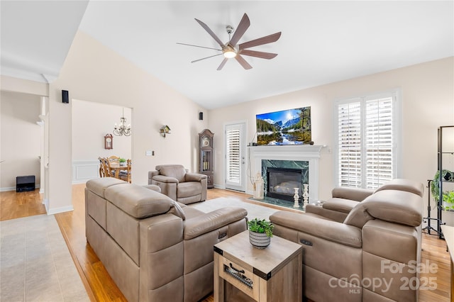 living area featuring lofted ceiling, light wood-style flooring, a premium fireplace, baseboards, and ceiling fan with notable chandelier