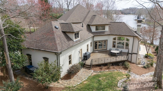 rear view of property with stucco siding, roof with shingles, a chimney, and a deck with water view