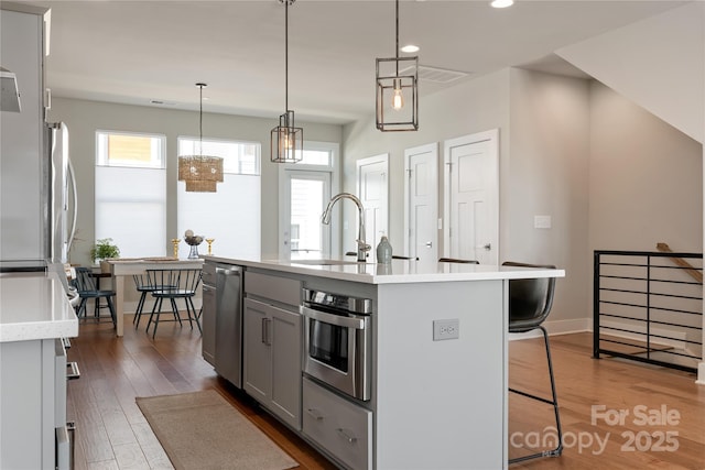 kitchen with dark wood-type flooring, gray cabinets, appliances with stainless steel finishes, a kitchen island with sink, and hanging light fixtures