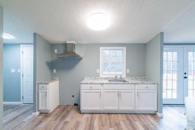 kitchen with wall chimney range hood, sink, light stone counters, white cabinets, and light wood-type flooring