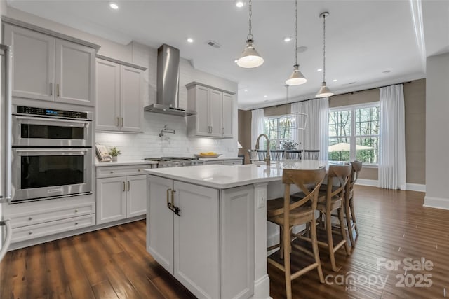kitchen featuring an island with sink, stainless steel appliances, light countertops, wall chimney exhaust hood, and dark wood-style floors