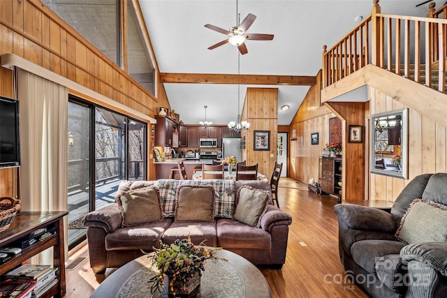 living room with ceiling fan with notable chandelier, high vaulted ceiling, wooden walls, beamed ceiling, and light hardwood / wood-style floors