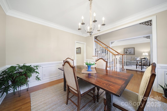 dining area featuring wainscoting, ornamental molding, wood finished floors, stairs, and a notable chandelier