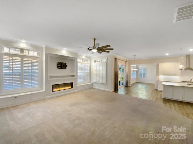 unfurnished living room with ceiling fan with notable chandelier, light carpet, and ornamental molding