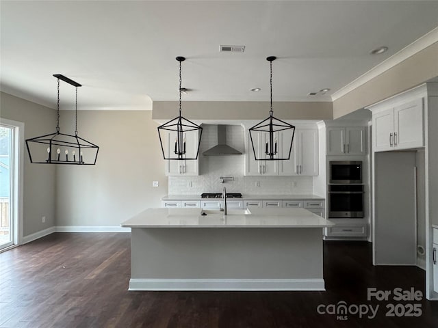 kitchen featuring decorative light fixtures, white cabinets, a kitchen island with sink, stainless steel oven, and wall chimney range hood