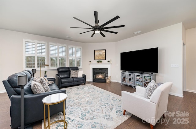 living area featuring dark wood-style flooring, visible vents, a ceiling fan, a glass covered fireplace, and baseboards