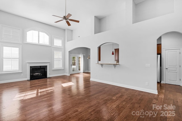 unfurnished living room with a ceiling fan, a high ceiling, wood finished floors, arched walkways, and a glass covered fireplace