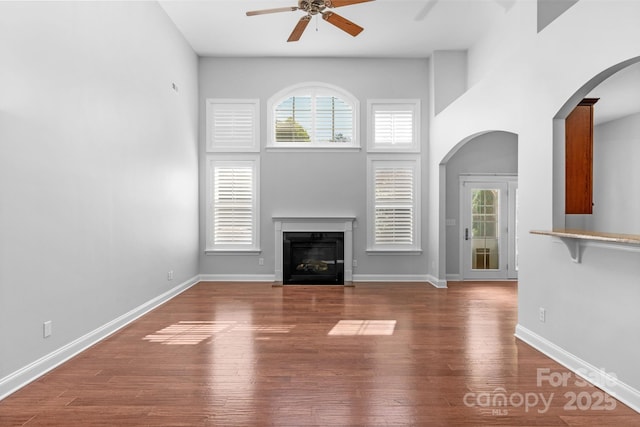 unfurnished living room featuring a ceiling fan, arched walkways, a glass covered fireplace, wood finished floors, and a high ceiling