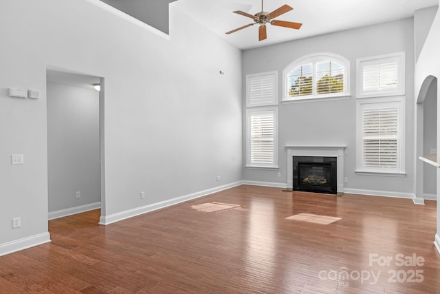 unfurnished living room with a ceiling fan, a towering ceiling, a glass covered fireplace, wood finished floors, and baseboards