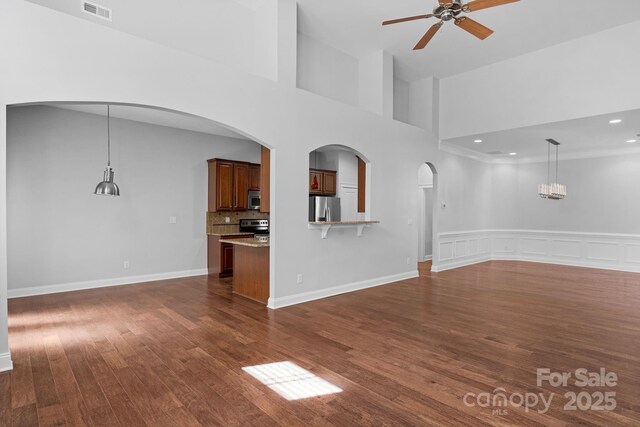 unfurnished living room with ceiling fan, arched walkways, dark wood-style floors, a towering ceiling, and a wainscoted wall